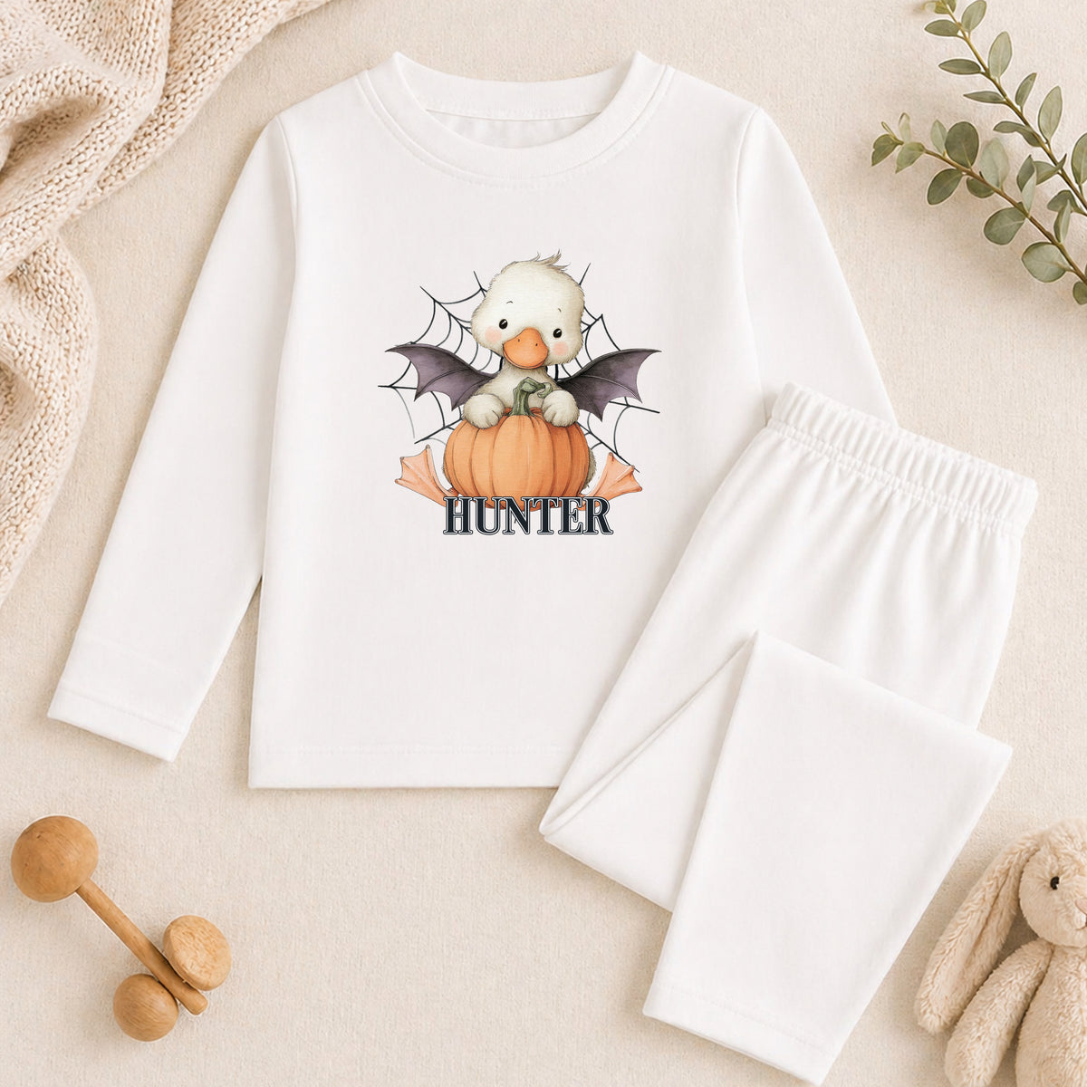 A white long-sleeved shirt with a cartoon image of a duck wearing a bat costume and holding a pumpkin, along with a pair of white pants and a stuffed animal.
