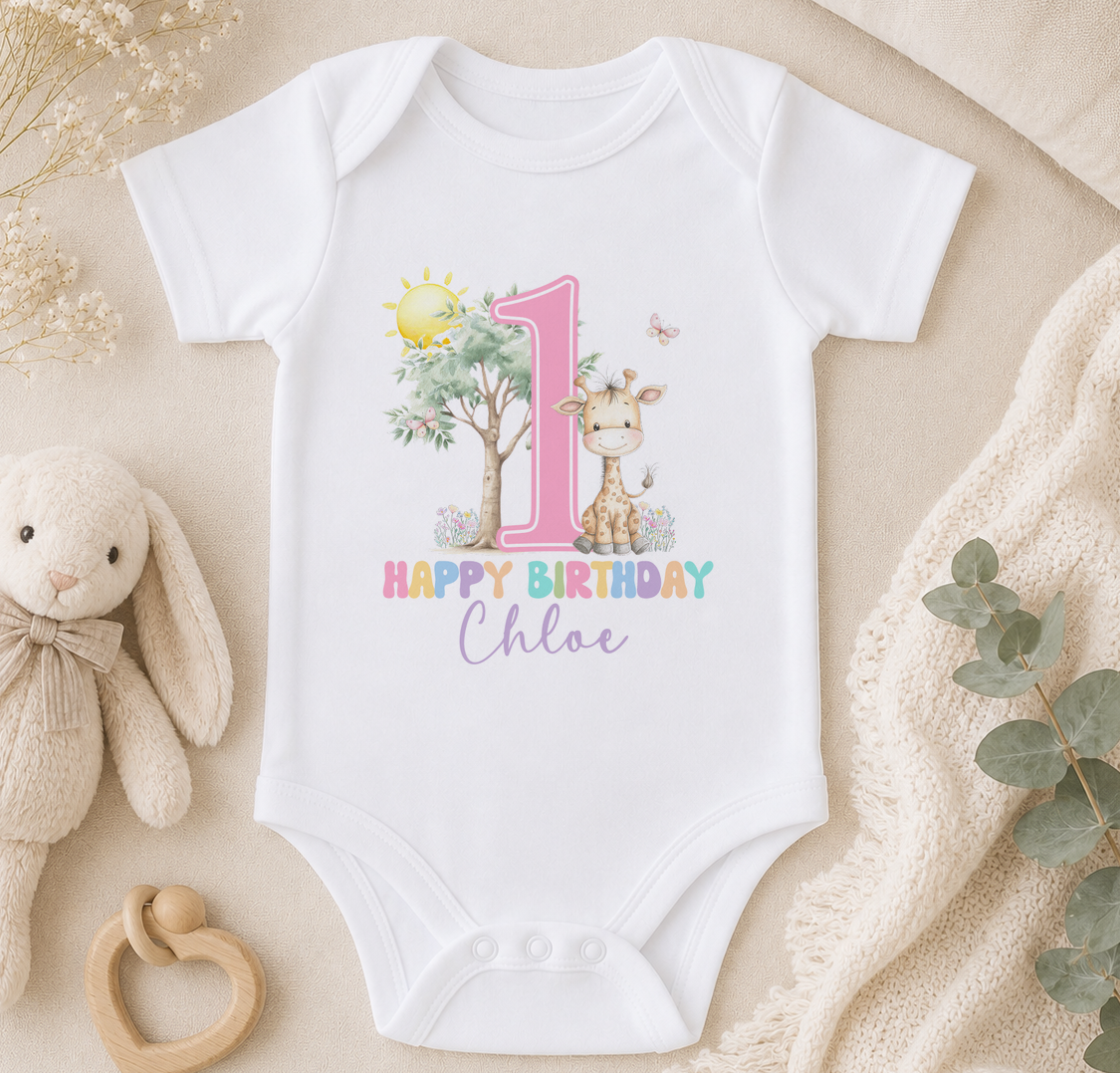 A white onesie with a giraffe and the number 1, along with the text "Happy Birthday Chloe" printed on it, is displayed on a bed surrounded by stuffed animals and greenery.