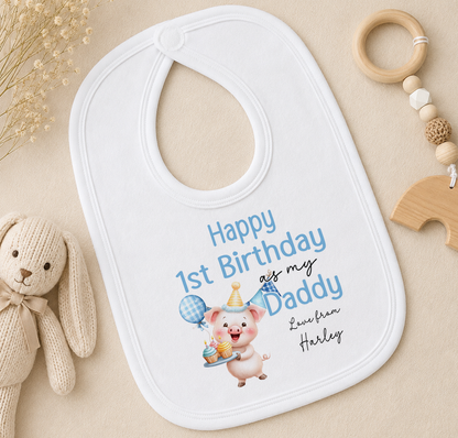 A white baby bib with a personalized message, a cute cartoon pig wearing a party hat, and a wooden toy ring are displayed on a beige surface.