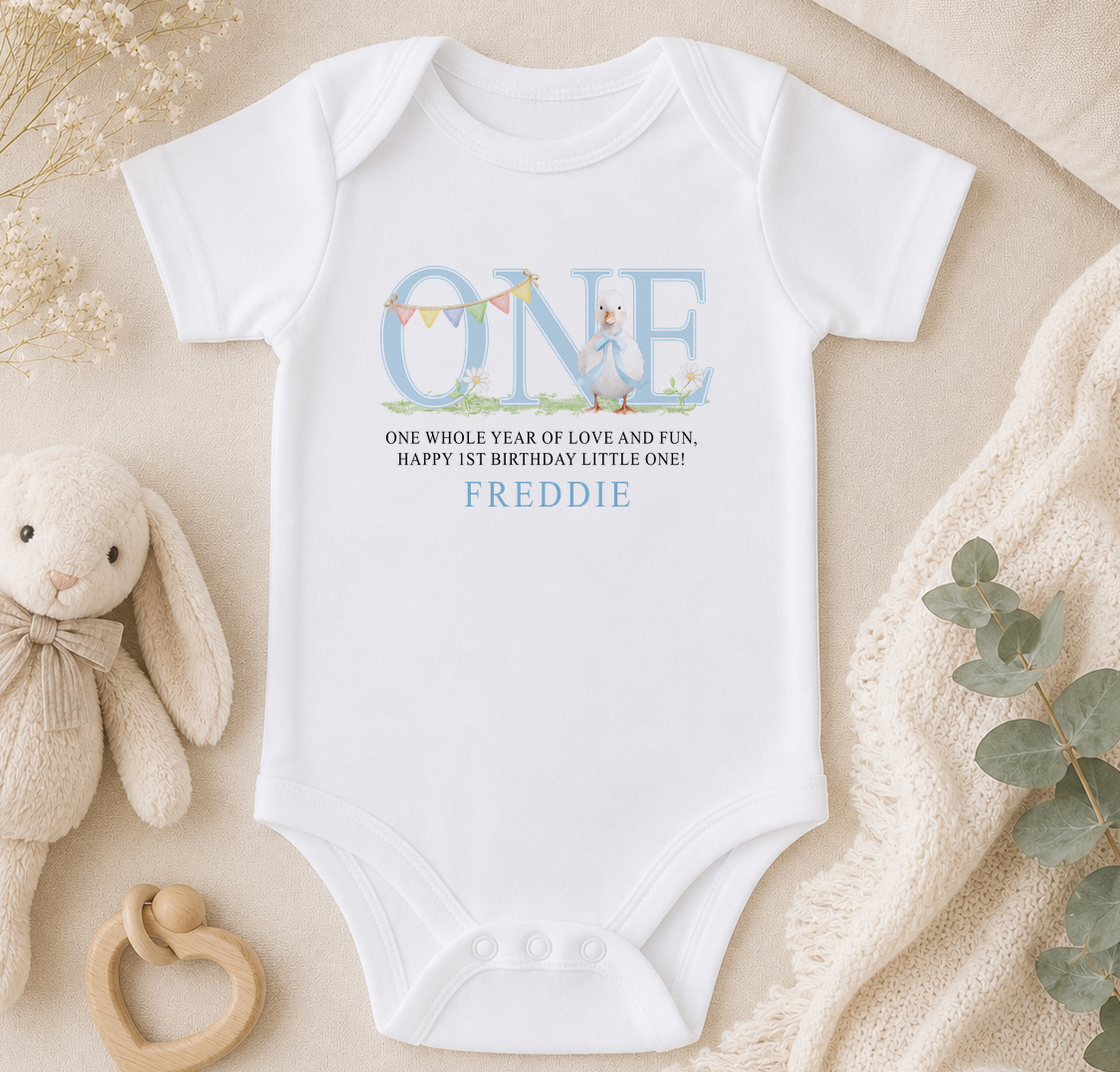 A white onesie with the text "ONE whole year of love and fun, happy 1st birthday little one!" printed on it, along with the name "FREDDIE". The onesie is placed on a beige surface, surrounded by a stuffed bunny, a wooden bead rattle, and some greenery.