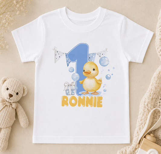 A white t-shirt with a cartoon duck and the number 1, along with the text "RONNIE" printed on it, is displayed on a beige surface.