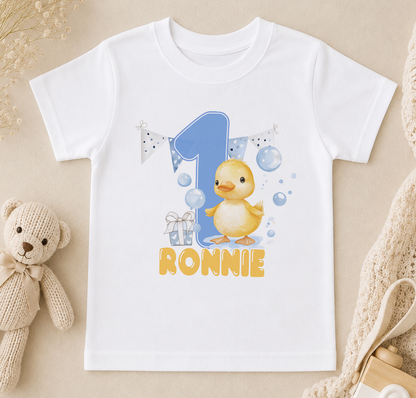 A white t-shirt with a cartoon duck and the number 1, along with the text "RONNIE" printed on it, is displayed on a beige surface.