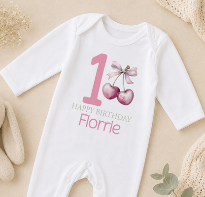A white onesie with a pink number 1 and a bow, along with the text "Happy Birthday Florrie" printed on it, is displayed on a beige surface.