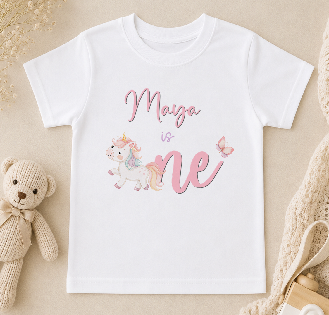 A white t-shirt with a printed design of a unicorn and the text "Maya is one" printed on it.