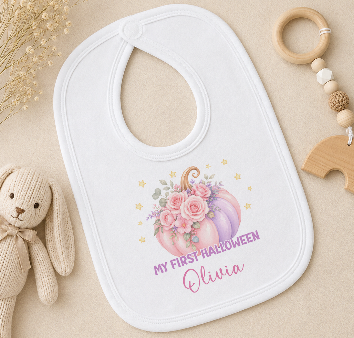 A white baby bib with a pumpkin and flowers design, along with a wooden toy and a wooden bead ring, all arranged on a beige surface.