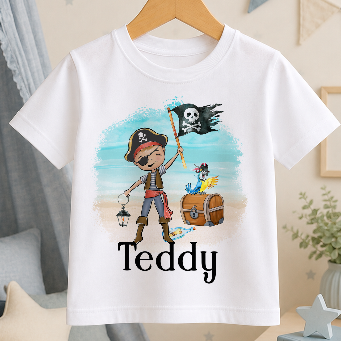 A white t-shirt with a pirate-themed graphic design, including a pirate character, a pirate flag, and a bird. The word "Teddy" is printed below the design.