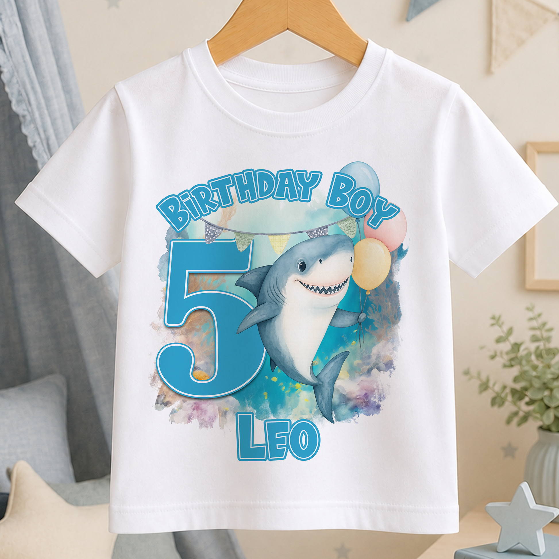 A white t-shirt with a graphic design featuring a shark and balloons, along with the text "Birthday Boy Leo" printed on it.