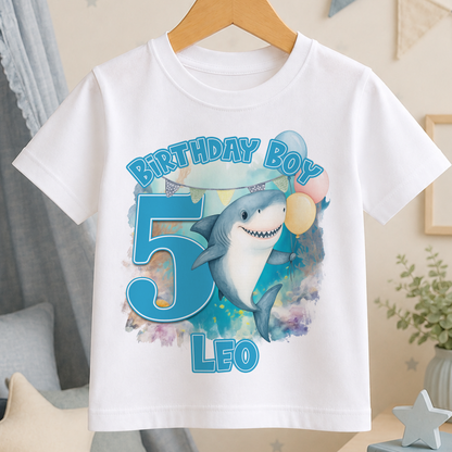 A white t-shirt with a graphic design featuring a shark and balloons, along with the text "Birthday Boy Leo" printed on it.