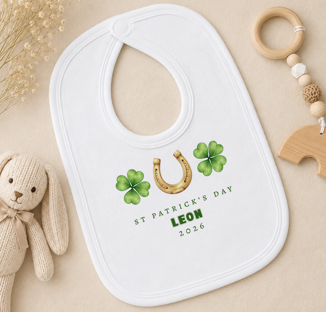 A white baby bib with a St. Patrick's Day design, featuring a horseshoe and four-leaf clovers, is placed on a beige surface alongside a stuffed rabbit and a wooden bead ringer.