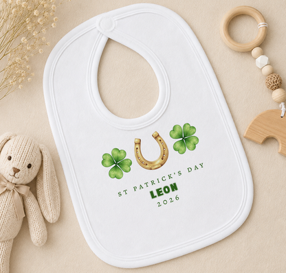 A white baby bib with a St. Patrick's Day design, featuring a horseshoe and four-leaf clovers, is placed on a beige surface alongside a stuffed rabbit and a wooden bead ringer.
