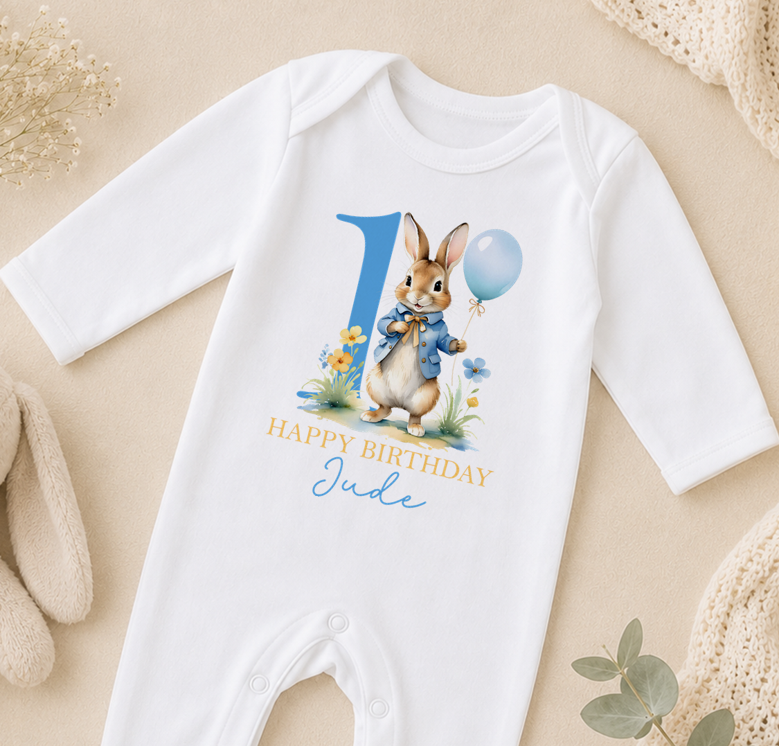A white onesie with a bunny character and a blue balloon, along with the text "Happy Birthday Jude" printed on it.