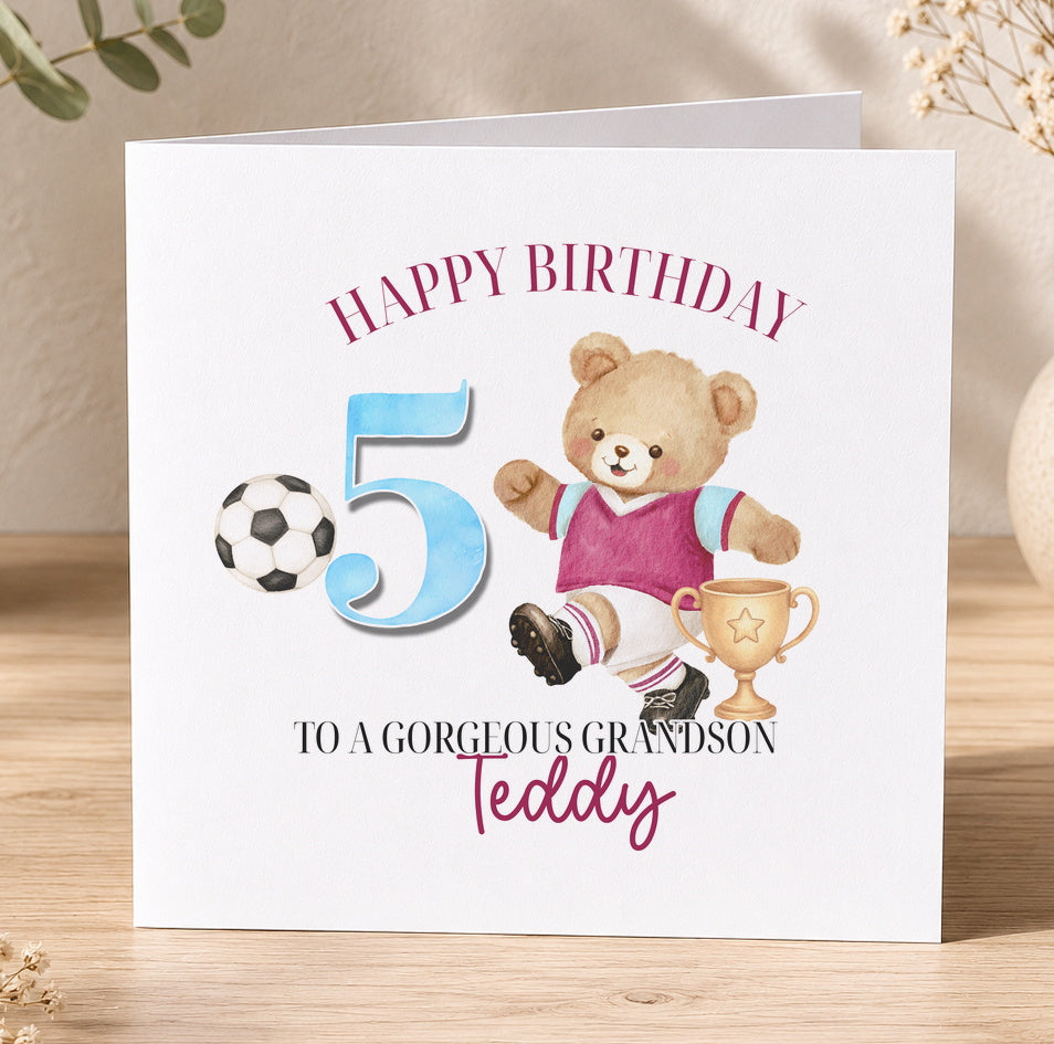 Square birthday card standing on a wooden surface, reading 'Happy Birthday 5, To A Gorgeous Grandson, Teddy', illustrated with a teddy bear in a football kit kicking a ball beside a gold trophy