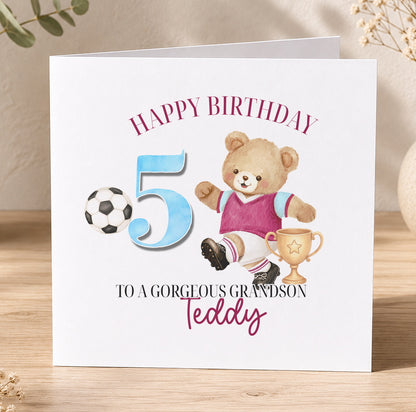 Square birthday card standing on a wooden surface, reading 'Happy Birthday 5, To A Gorgeous Grandson, Teddy', illustrated with a teddy bear in a football kit kicking a ball beside a gold trophy