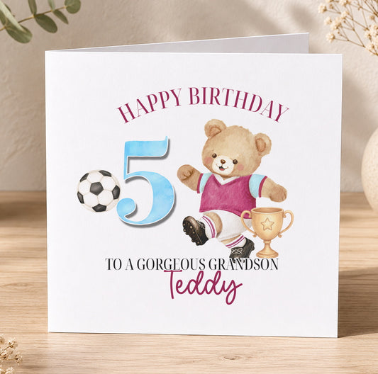 Square birthday card standing on a wooden surface, reading 'Happy Birthday 5, To A Gorgeous Grandson, Teddy', illustrated with a teddy bear in a football kit kicking a ball beside a gold trophy