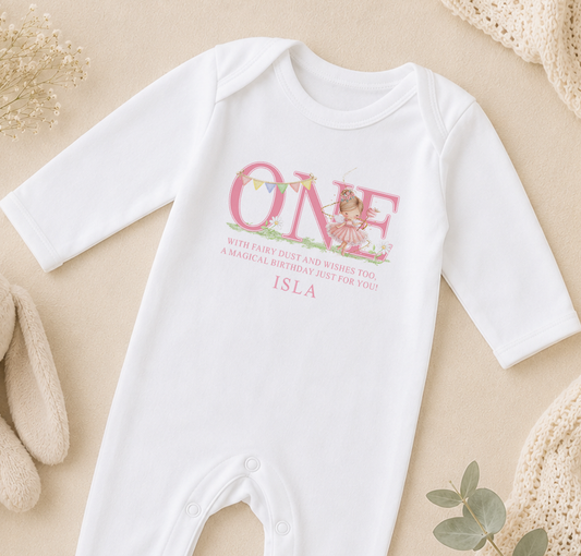 A white onesie with a personalized text and a fairy image printed on it, placed on a beige surface with some decorative elements in the background.