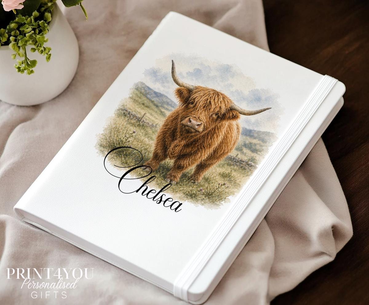 White A5 notebook with a PU leather cover and elastic closure band, printed with a watercolour Highland cow illustration and personalised with the name Chelsea in script, photographed on a beige throw beside a vase of flowers