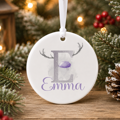 A white oval ornament with the letter "E" and the name "Emma" printed on it, hanging on a Christmas tree.