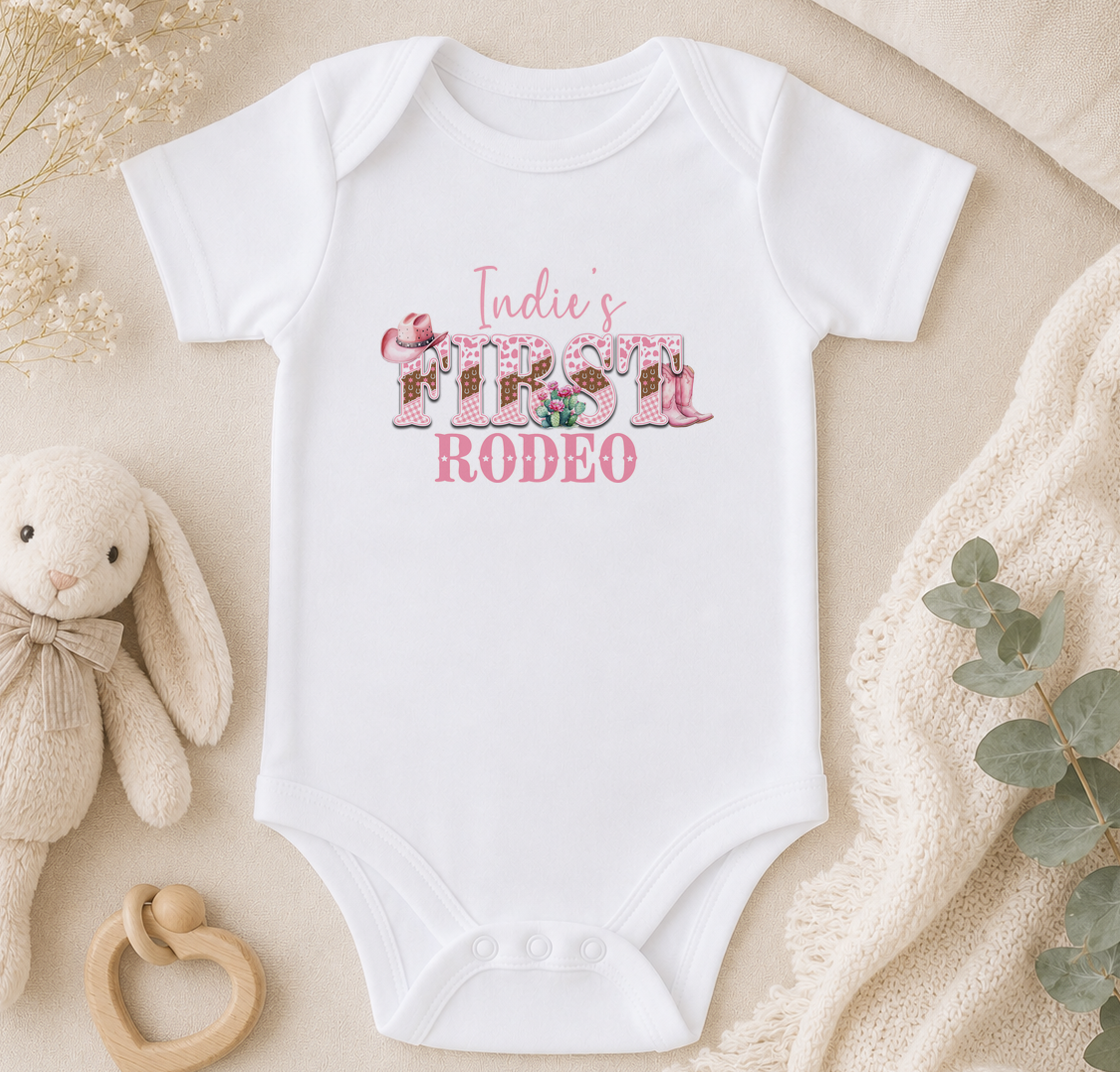A white baby onesie with the text "Indie's First Rodeo" printed on it, along with a stuffed animal and some greenery.
