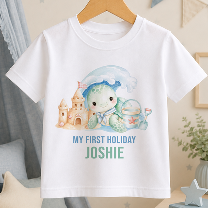 A white t-shirt with a printed design of a turtle and a castle, along with the text "My first holiday Joshie" printed on it.