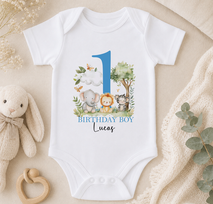 A white onesie with a blue number 1 and a design of animals, including an elephant, lion, and zebra, is displayed on a beige surface.