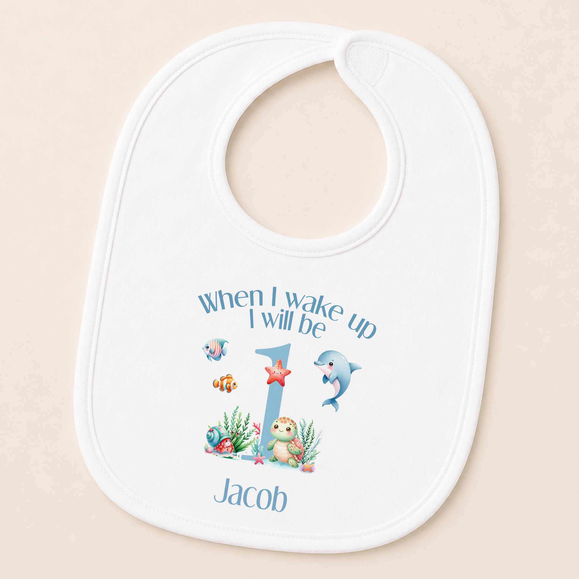 A white baby bib with a personalized design featuring a cartoon image of a dolphin and the text "When I wake up I will be Jacob" along with a number 1.
