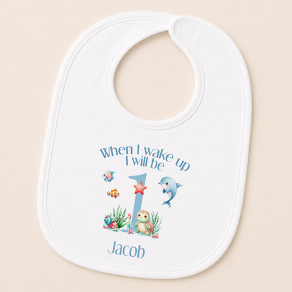 A white baby bib with a personalized design featuring a cartoon image of a dolphin and the text "When I wake up I will be Jacob" along with a number 1.