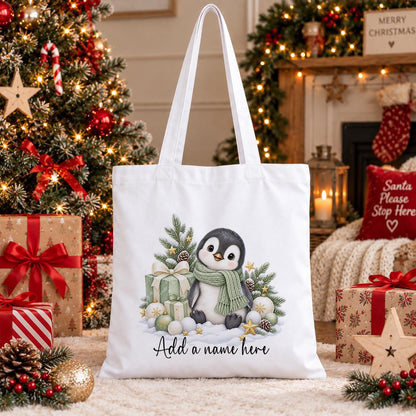 White cotton tote bag printed with a penguin in a sage green scarf beside wrapped gifts, pine and baubles, above a name in script.