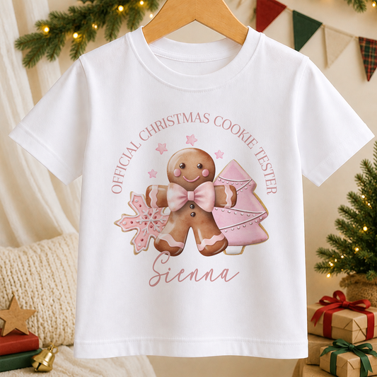A white t-shirt with a Christmas-themed graphic design featuring a gingerbread man, a snowflake, and a pink bow.