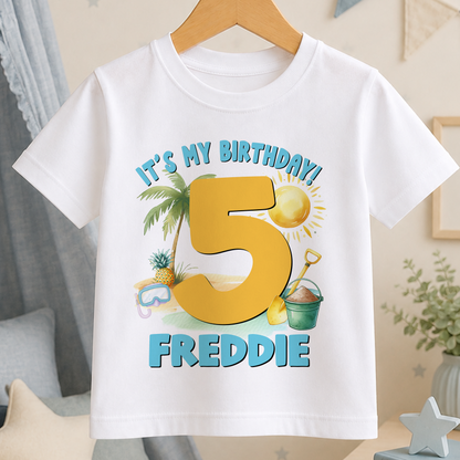 A white t-shirt with a graphic design featuring a large number 5, a palm tree, a beach scene, and a bucket, along with the text "It's my birthday! FREDDIE".
