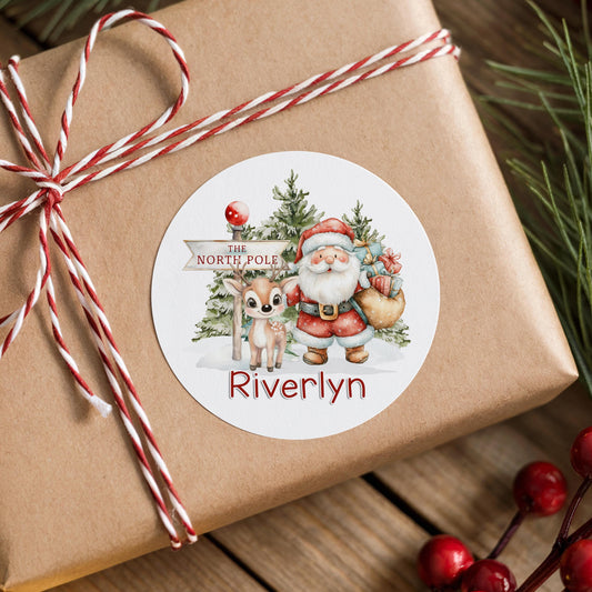 A brown gift box wrapped with red and white twine, featuring a round sticker with a Santa Claus and reindeer illustration and the name "Riverlyn" written on it.