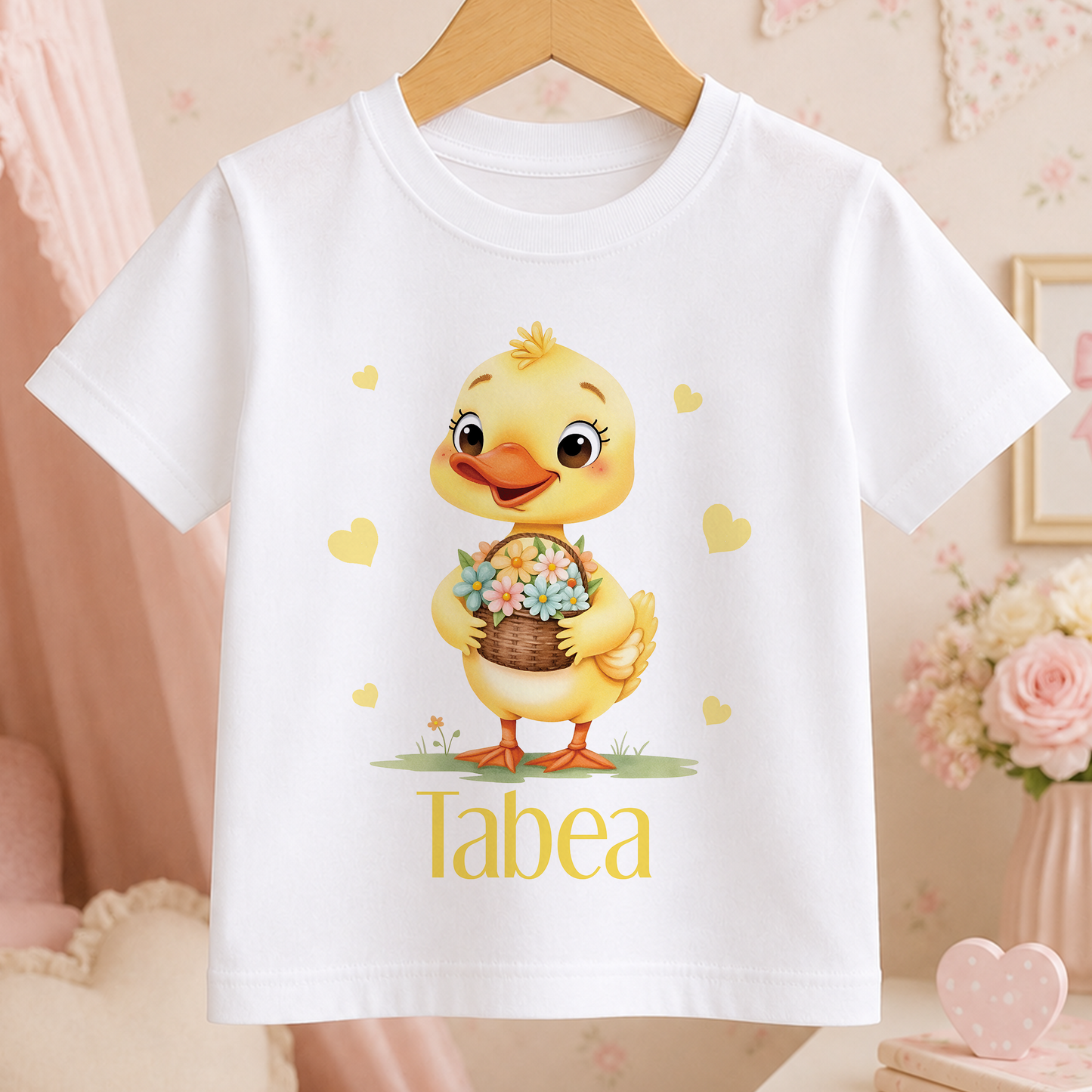 A white t-shirt with a cartoon image of a yellow duck holding a basket of flowers, surrounded by hearts. The word "Tabea" is written below the duck.