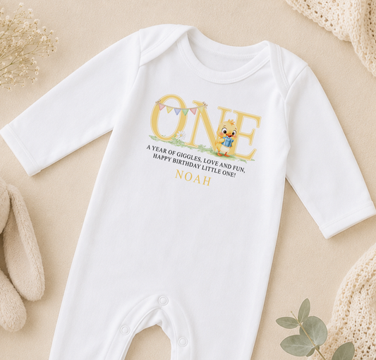 A white onesie with a personalized message and a cartoon character printed on it, placed on a beige surface with some decorative elements in the background.