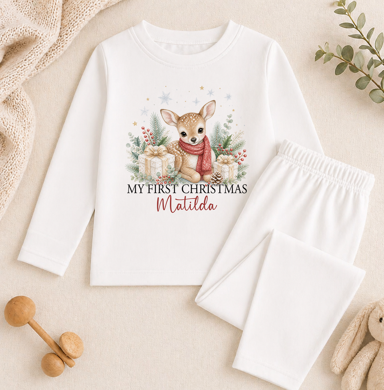 A white long-sleeved shirt with a printed image of a deer and Christmas decorations, along with a pair of white pajama pants, and some stuffed animals.