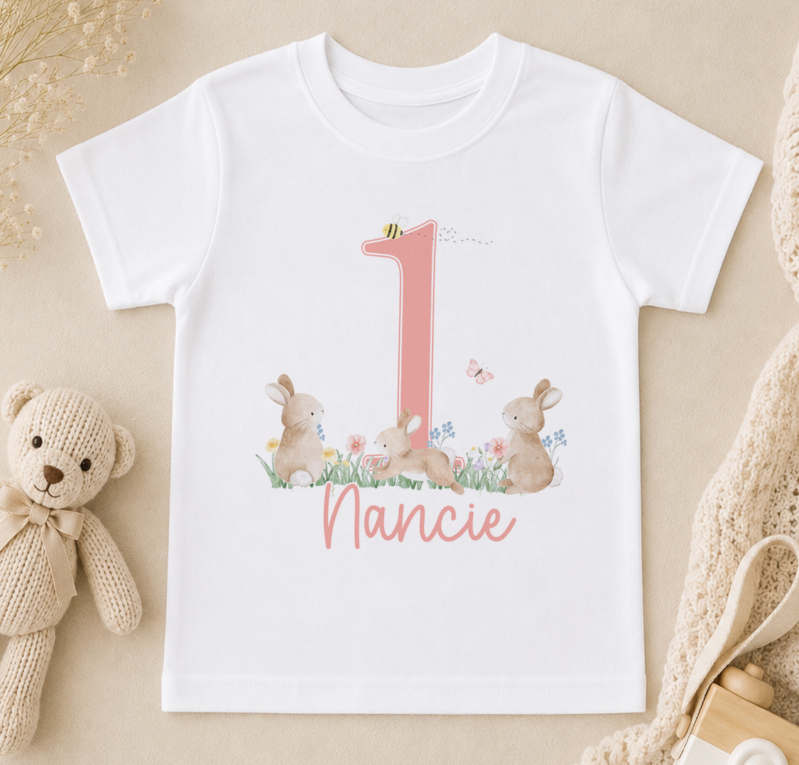 A white t-shirt with a pink number one and a bunny illustration, along with the name "Nancie" written below.