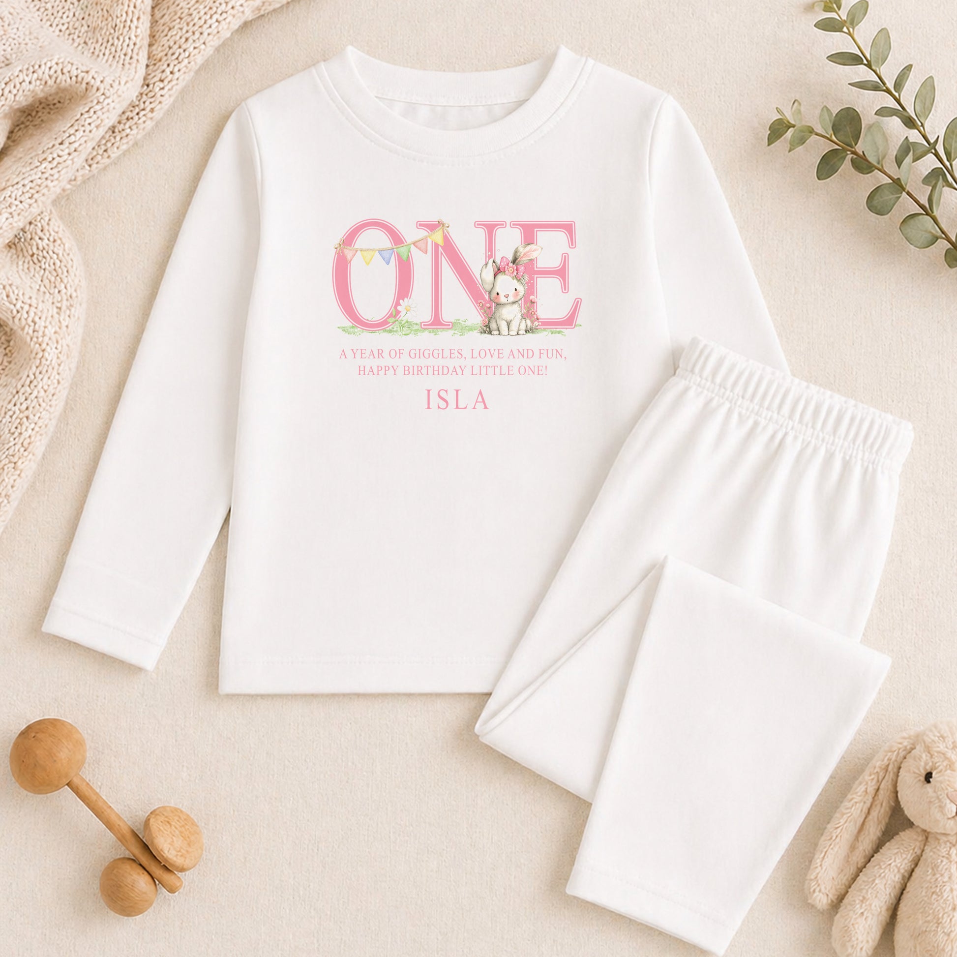 A white long-sleeved shirt with a pink and white design, featuring the text "ONE" and a small illustration of a cat, along with the text "A year of giggle, love and fun! Happy birthday little one! ISLA". The shirt is laid out on a beige surface, accompanied by a pair of white pajama pants and a stuffed animal.