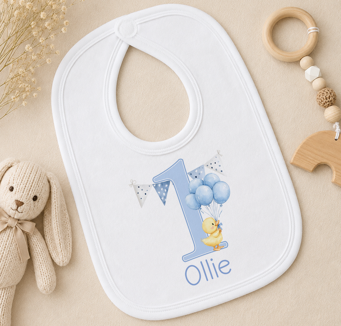 A white bib with a personalized number 1 and a yellow duck wearing a blue bow tie, along with a wooden toy and some decorative elements.