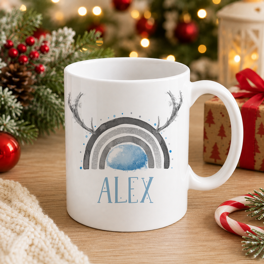 A white mug with a graphic design featuring a rainbow and antlers, along with the name "ALEX" printed on it, sits on a wooden surface surrounded by holiday decorations such as a pine tree, ornaments, and a wrapped gift.
