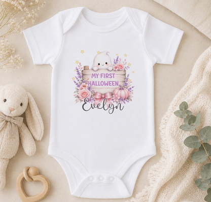 A white baby onesie with a Halloween-themed design, including a ghost, flowers, and a sign that says "My first Halloween Evelyn". The onesie is placed on a beige surface, surrounded by a stuffed bunny, a wooden toy, and some greenery.
