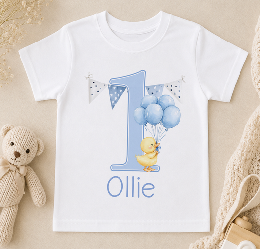 A white t-shirt with a blue number one and a yellow duck holding blue balloons, along with the name "Ollie" written on it.