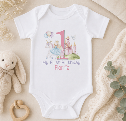 A white onesie with a pink number 1 and a castle illustration, along with a stuffed bunny and a baby rattle, all arranged on a beige surface.