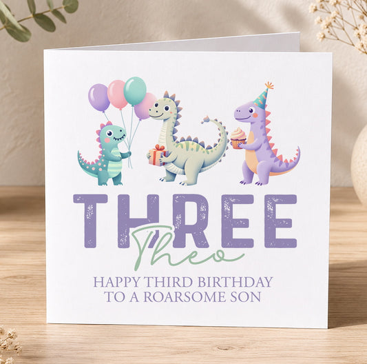 Third birthday card illustrated with three dinosaurs and balloons, printed with 'Three' and 'Happy third birthday to a roaring son' as a personalisation example.