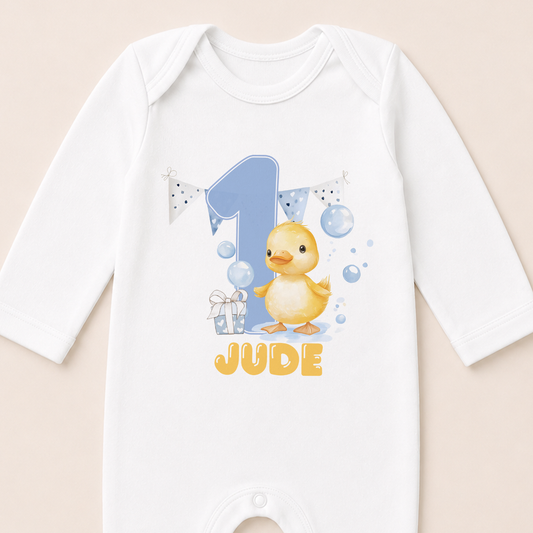 A white onesie with a cartoon image of a yellow duck and the text "JUDE" printed on it.