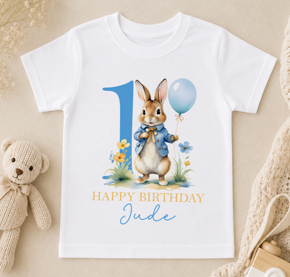 White personalised t-shirt printed with a bunny holding a blue balloon surrounded by flowers and the message Happy Birthday Jude.