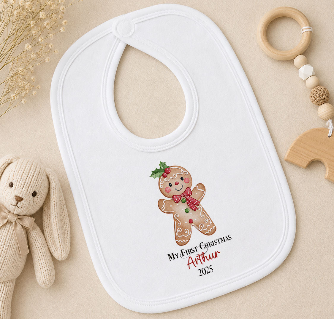 A white baby bib with a gingerbread man design and the text "My First Christmas Arthur" printed on it, along with a wooden toy and a wooden bead ring.