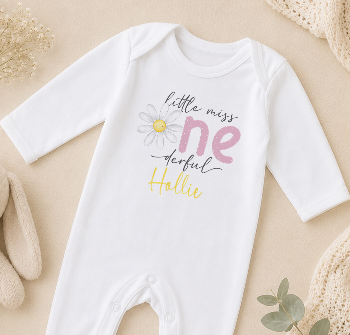 A white onesie with a flower design and text printed on it, laid out on a beige surface.