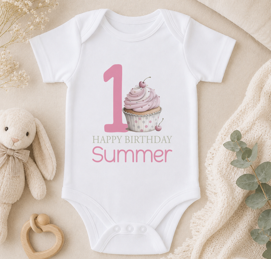 A white onesie with a pink cupcake and the text "Happy Birthday Summer" printed on it, along with a number "1" and a teddy bear.