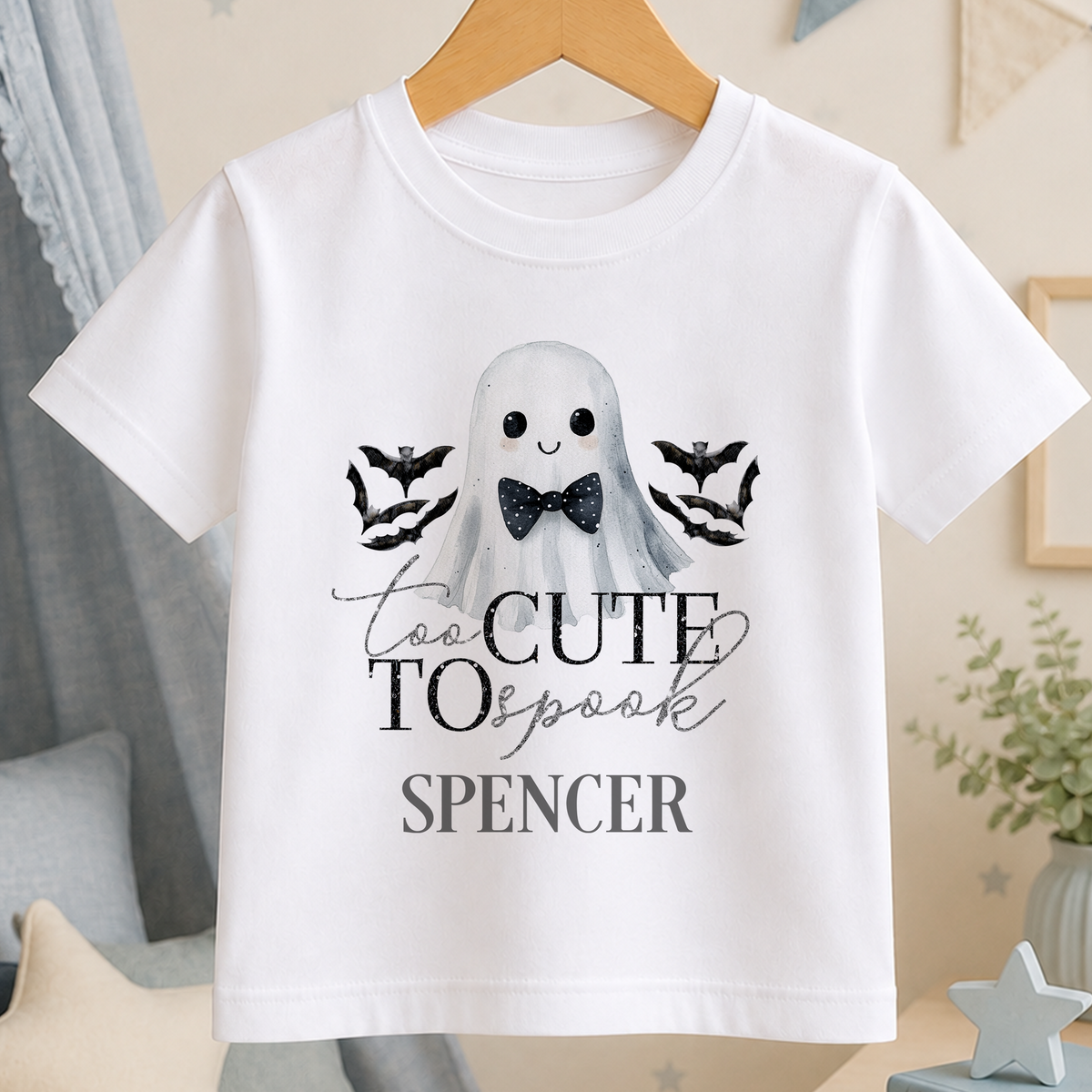 A white t-shirt with a graphic design featuring a ghost character and bats, along with the text "Too Cute to Spook" and "Spenser" printed on it.