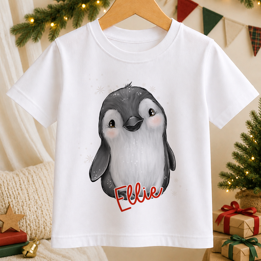 A white t-shirt with a cartoon penguin design and the name "Ellie" written on it, hanging on a wooden hanger.