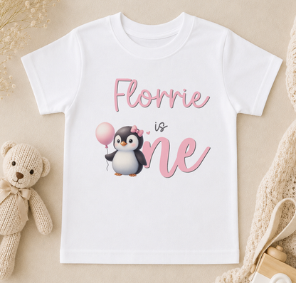 A white t-shirt with a pink penguin and the text "Flourrie is one" printed on it, along with a teddy bear and other baby items.