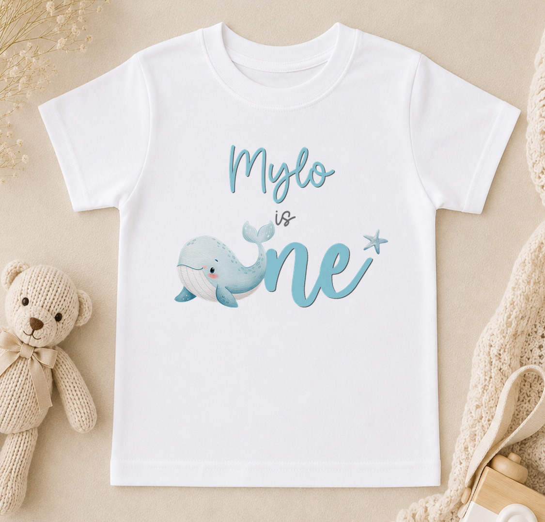 A white t-shirt with a blue whale graphic and the text "Mylo is one" printed on it, along with a teddy bear and other baby items.